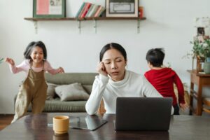 A tired mother working on a laptop while her children play around indoors, highlighting remote work challenges.