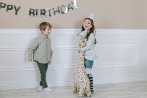 Two smiling children at a birthday party indoors, wearing party hats and holding a stuffed toy giraffe.