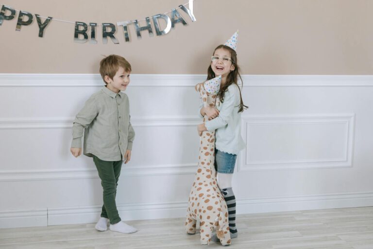 Two smiling children at a birthday party indoors, wearing party hats and holding a stuffed toy giraffe.