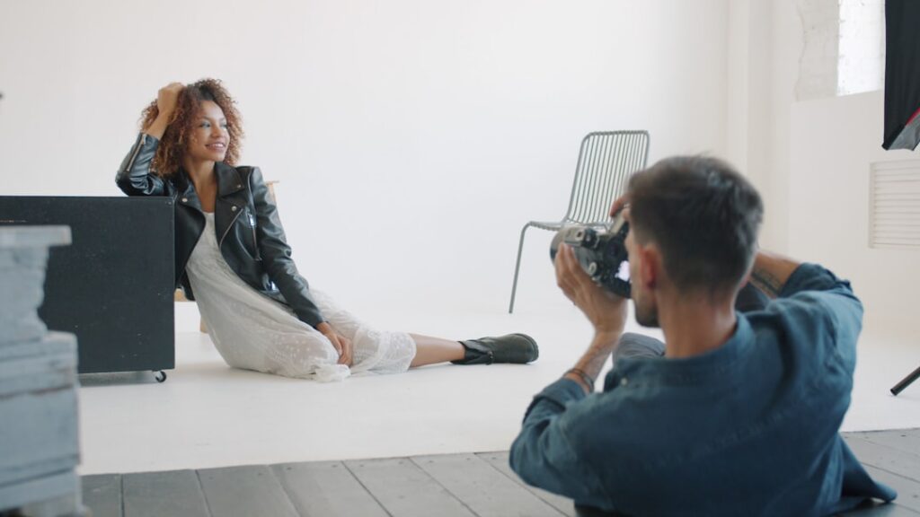 Sexy girl in fashionable clothing is posing for camera in studio working with professional photographer sitting on floor on white background. People and lifestyle concept.