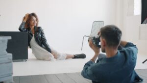 Sexy girl in fashionable clothing is posing for camera in studio working with professional photographer sitting on floor on white background. People and lifestyle concept.