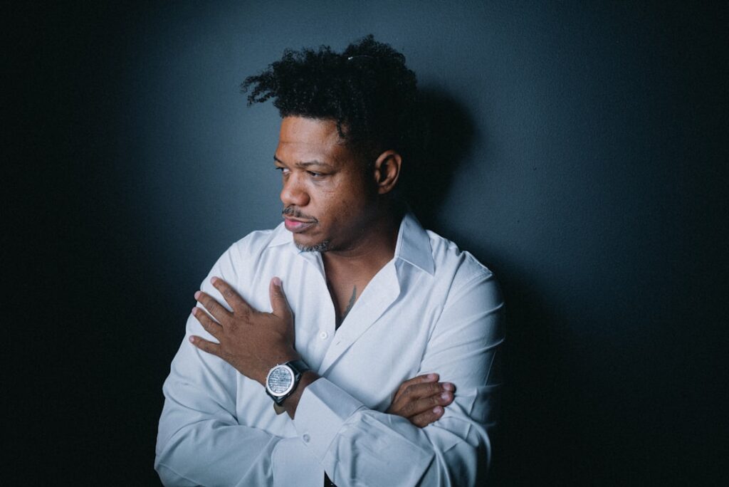 A thoughtful black man in a crisp white shirt poses with arms crossed, gazing off-camera with a contemplative expression. The dark background highlights his presence, creating a sense of introspection and quiet confidence