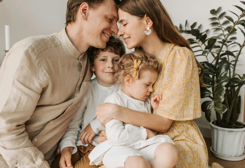 Photo by Vlada Karpovich A loving family of four shares a tender moment in a cozy indoor setting, embracing and smiling.